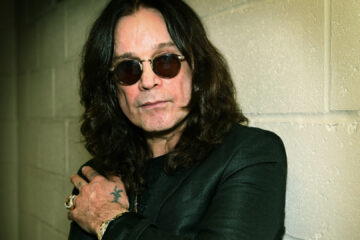 Ozzy Osbourne com óculos escuros, em frente a uma parede de tijolos, símbolo da campanha para nomear o aeroporto de Birmingham em sua homenagem.