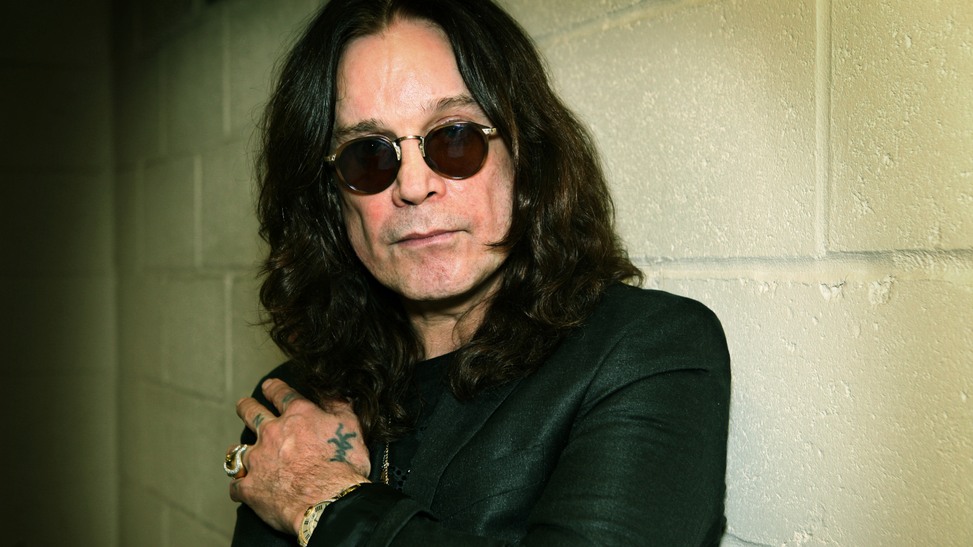 Ozzy Osbourne com óculos escuros, em frente a uma parede de tijolos, símbolo da campanha para nomear o aeroporto de Birmingham em sua homenagem.