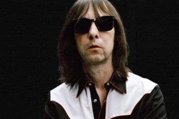 Bobby Gillespie, vocalista do Primal Scream, em foto promocional vestindo camisa preta com detalhes brancos, óculos escuros e fundo preto – Crédito: Adam Peter Johnson