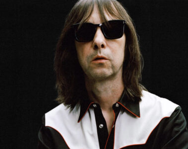Bobby Gillespie, vocalista do Primal Scream, em foto promocional vestindo camisa preta com detalhes brancos, óculos escuros e fundo preto – Crédito: Adam Peter Johnson