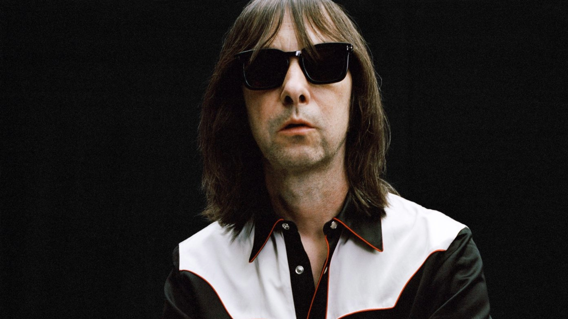 Bobby Gillespie, vocalista do Primal Scream, em foto promocional vestindo camisa preta com detalhes brancos, óculos escuros e fundo preto – Crédito: Adam Peter Johnson