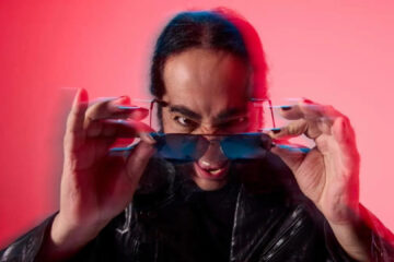 Daron Malakian, guitarrista do System of a Down e líder do Scars on Broadway, em ensaio promocional do álbum Addicted to the Violence, usando óculos escuros e jaqueta de couro, com fundo vermelho estilizado