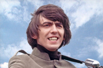 George Harrison sorrindo com o céu ao fundo durante os anos 60, segurando sua guitarra