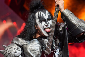 Gene Simmons com maquiagem clássica do Kiss tocando baixo e mostrando a língua durante um show ao vivo.