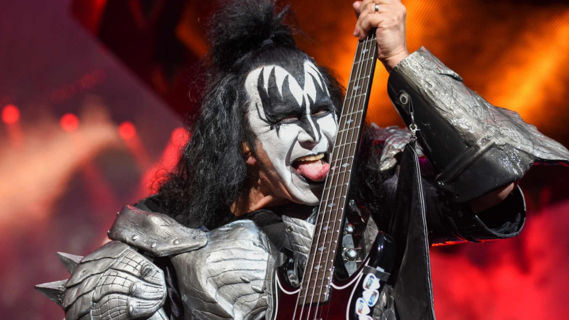 Gene Simmons com maquiagem clássica do Kiss tocando baixo e mostrando a língua durante um show ao vivo.
