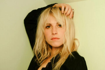 Retrato de Hayley Williams com expressão séria, cabelo loiro e blusa preta, em frente a uma parede verde-clara