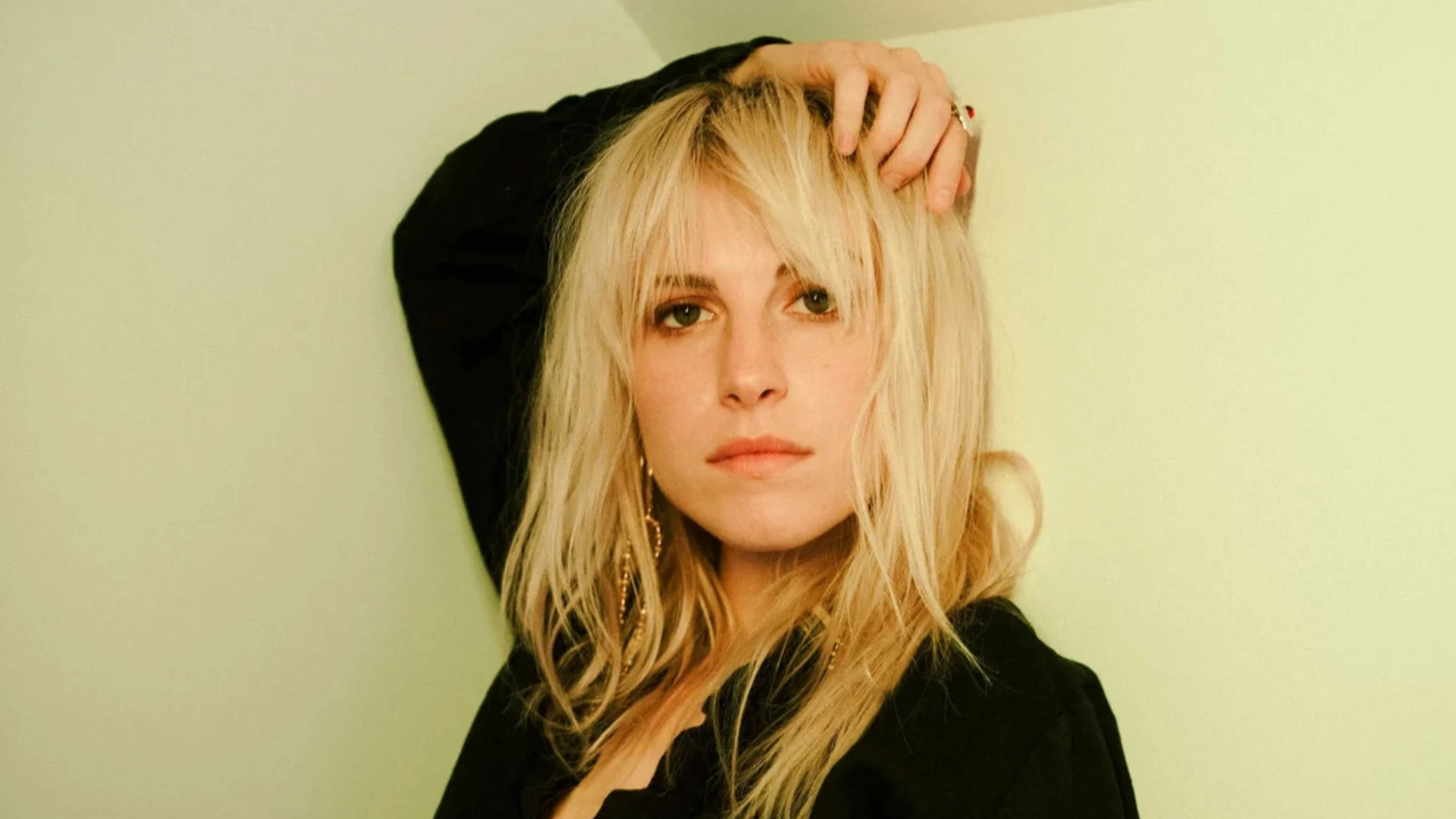 Retrato de Hayley Williams com expressão séria, cabelo loiro e blusa preta, em frente a uma parede verde-clara