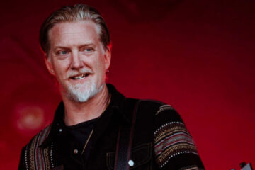 Josh Homme em apresentação ao vivo, sorrindo diante de fundo vermelho