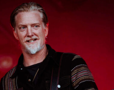 Josh Homme em apresentação ao vivo, sorrindo diante de fundo vermelho