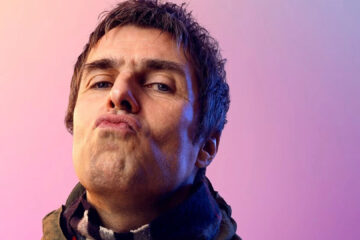 Retrato de Liam Gallagher encarando a câmera com expressão desafiadora, sobre fundo rosa e roxo.