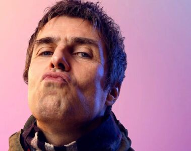 Retrato de Liam Gallagher encarando a câmera com expressão desafiadora, sobre fundo rosa e roxo.