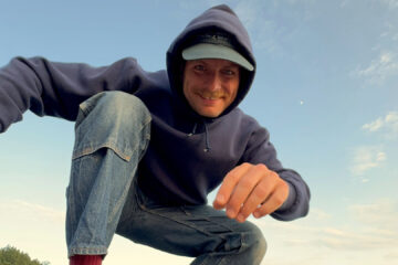 Mac DeMarco agachado em rocha à beira-mar, sorrindo, em clima informal — imagem usada na divulgação do show no Brasil em 2026