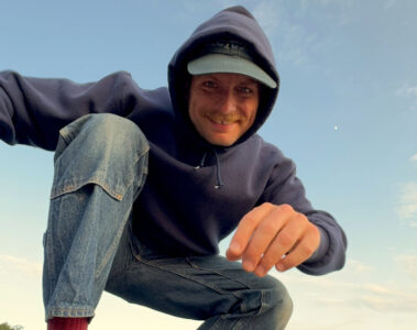 Mac DeMarco agachado em rocha à beira-mar, sorrindo, em clima informal — imagem usada na divulgação do show no Brasil em 2026