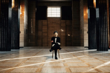 Artista PRESIDENT sentado em cadeira no centro de um salão escuro e imponente, com iluminação dramática