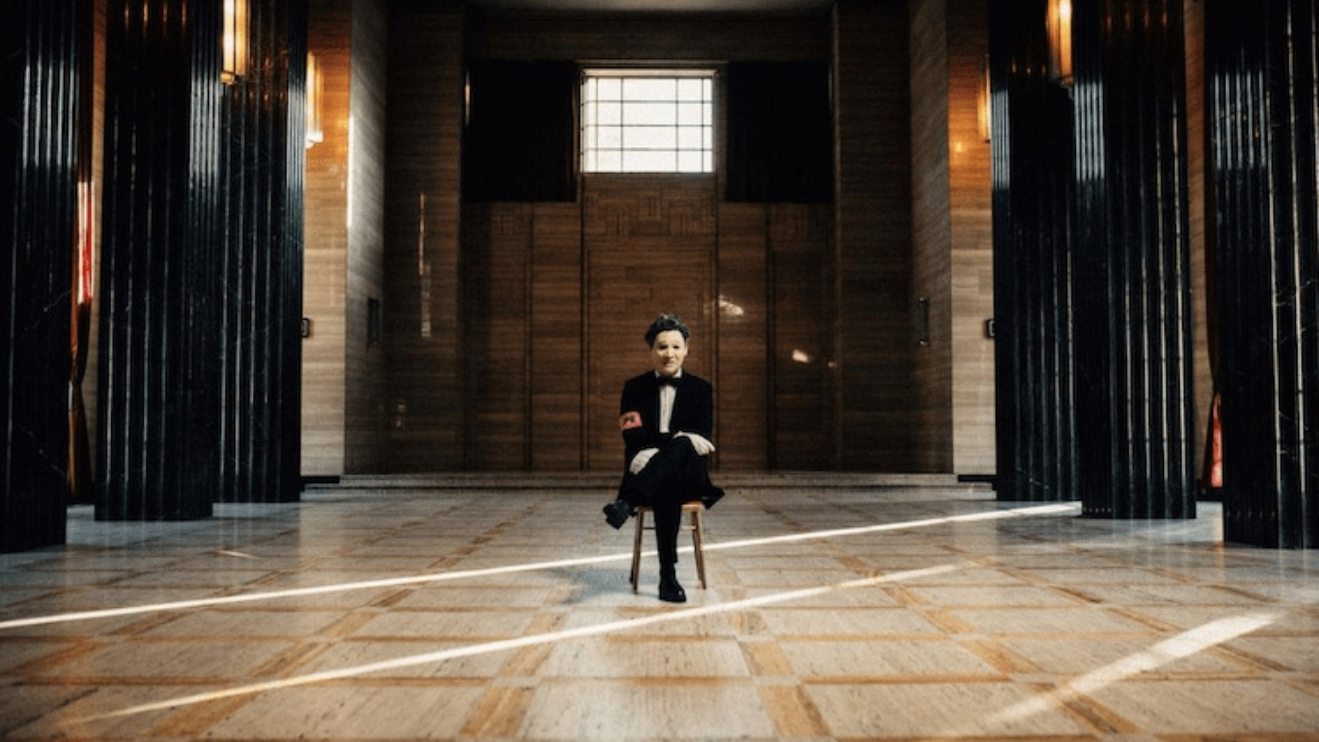 Artista PRESIDENT sentado em cadeira no centro de um salão escuro e imponente, com iluminação dramática