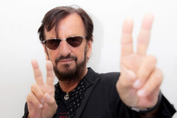 Ringo Starr durante apresentação ao vivo, segurando baquetas, com luz vermelha ao fundo