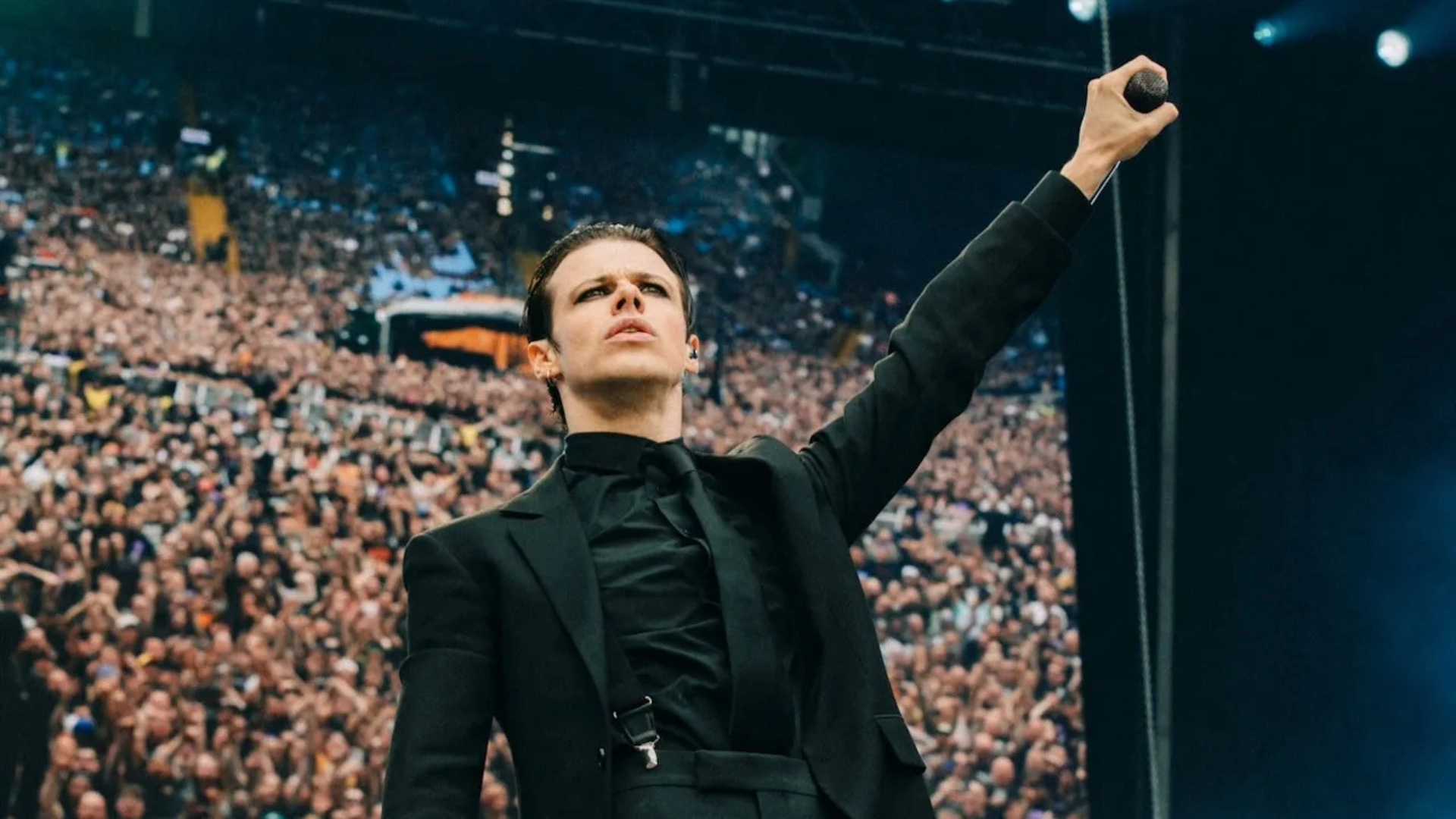 Yungblud com microfone erguido diante de uma multidão durante show ao ar livre