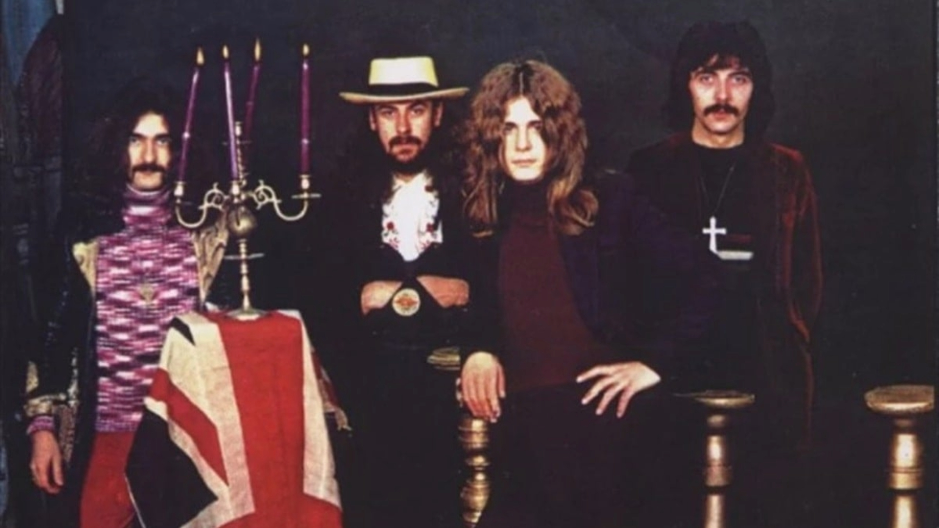 Foto clássica do Black Sabbath nos anos 1970, com Ozzy Osbourne, Tony Iommi, Geezer Butler e Bill Ward ao lado de um candelabro e bandeira britânica.