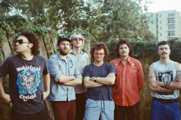 King Gizzard & The Lizard Wizard posa ao ar livre com os seis integrantes lado a lado, em clima descontraído.