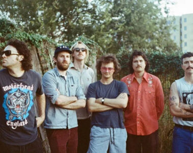 King Gizzard & The Lizard Wizard posa ao ar livre com os seis integrantes lado a lado, em clima descontraído.