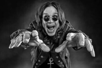 Ozzy Osbourne com os braços estendidos e expressão intensa, usando óculos escuros e anéis, em fundo escuro dramático