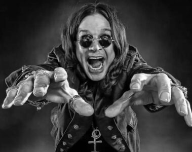 Ozzy Osbourne com os braços estendidos e expressão intensa, usando óculos escuros e anéis, em fundo escuro dramático