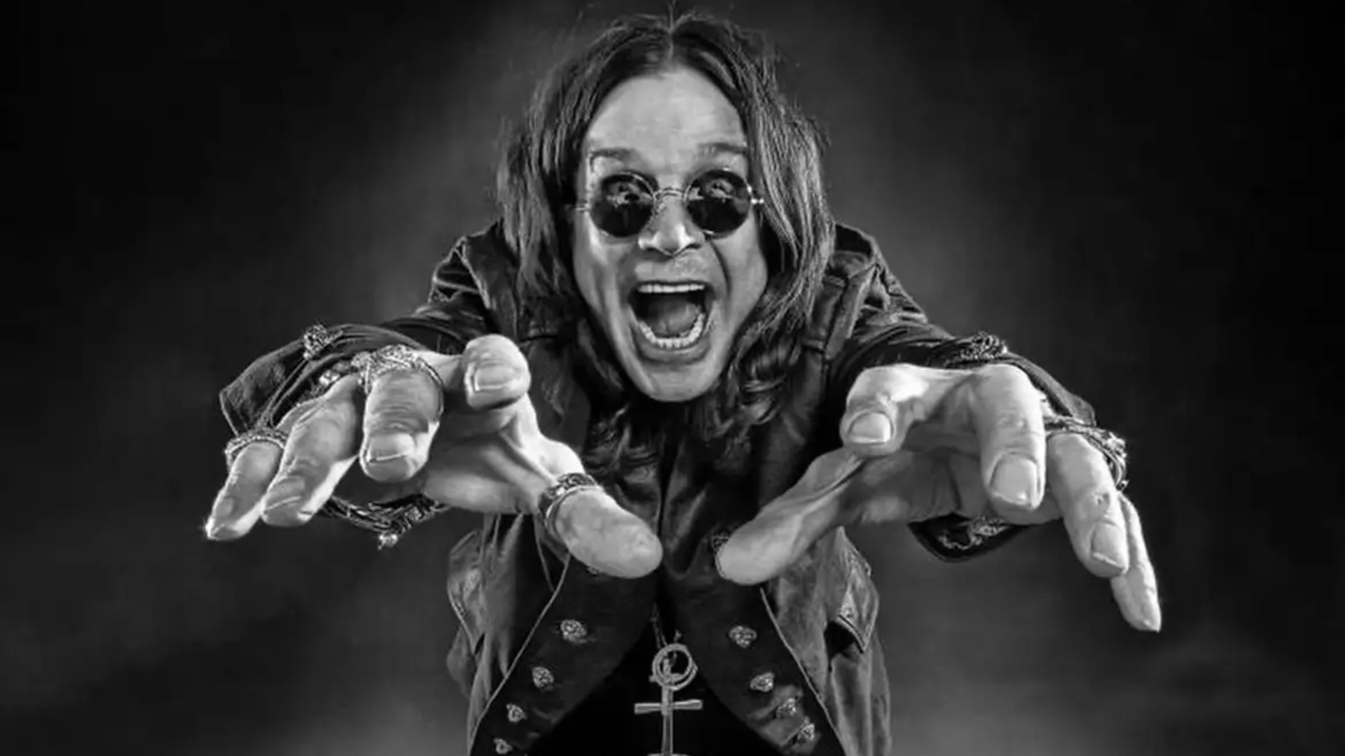 Ozzy Osbourne com os braços estendidos e expressão intensa, usando óculos escuros e anéis, em fundo escuro dramático