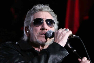 Roger Waters cantando ao vivo com óculos escuros e jaqueta de couro