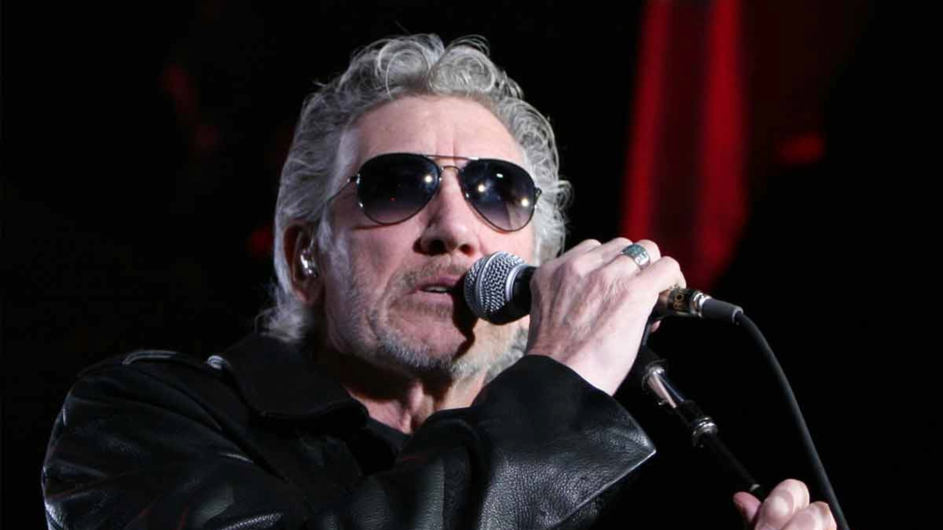 Roger Waters cantando ao vivo com óculos escuros e jaqueta de couro