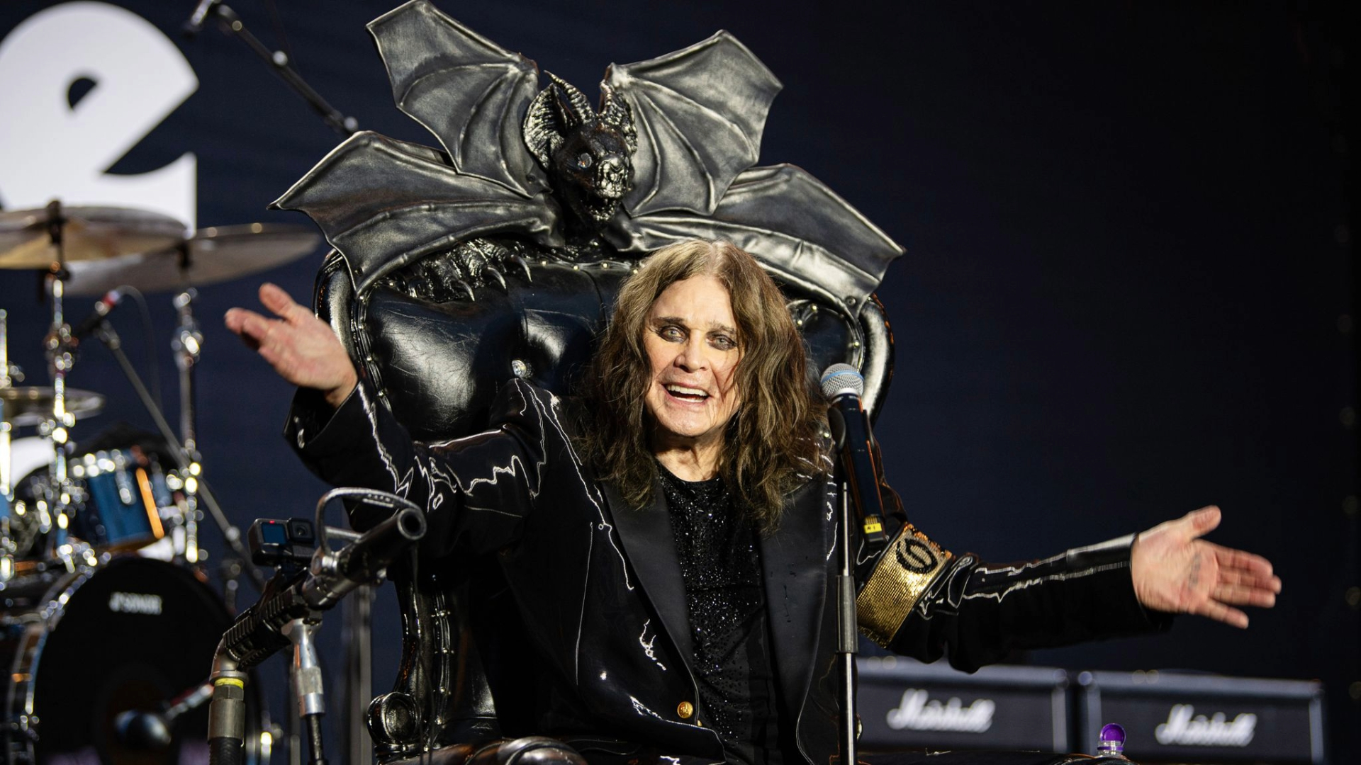 Ozzy Osbourne sorridente, usando jaqueta preta brilhante e braçadeira dourada com seu nome, se apresenta sentado em um trono durante o show de despedida do Black Sabbath em Birmingham. Foto de Ross Halfin.