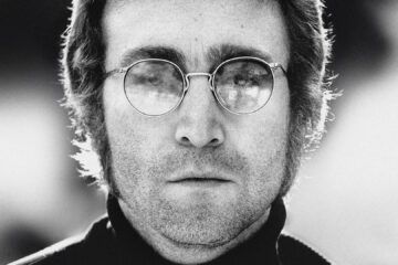 Retrato em preto e branco de John Lennon usando óculos redondos e expressão séria.