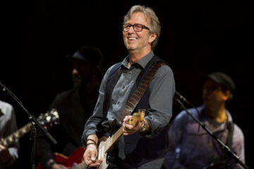 Eric Clapton sorrindo enquanto toca guitarra em um show, com músicos ao fundo e iluminação suave no palco.
