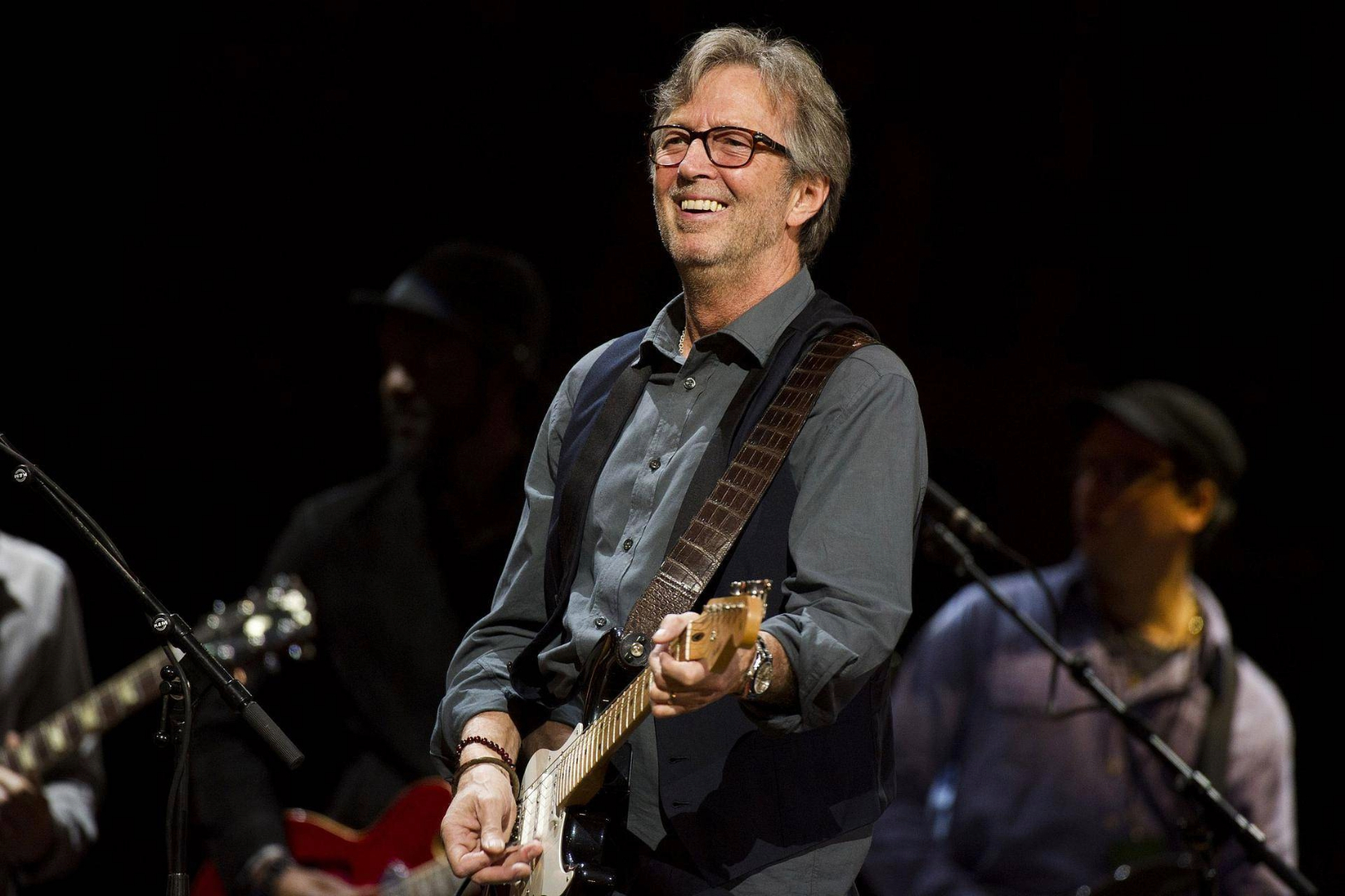 Eric Clapton sorrindo enquanto toca guitarra em um show, com músicos ao fundo e iluminação suave no palco.