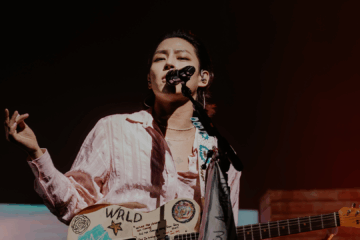 Woosung, vocalista do The Rose, cantando em um show ao vivo, segurando uma guitarra personalizada com desenhos e a inscrição WRLD, sob iluminação de palco.