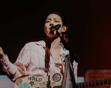 Woosung, vocalista do The Rose, cantando em um show ao vivo, segurando uma guitarra personalizada com desenhos e a inscrição WRLD, sob iluminação de palco.