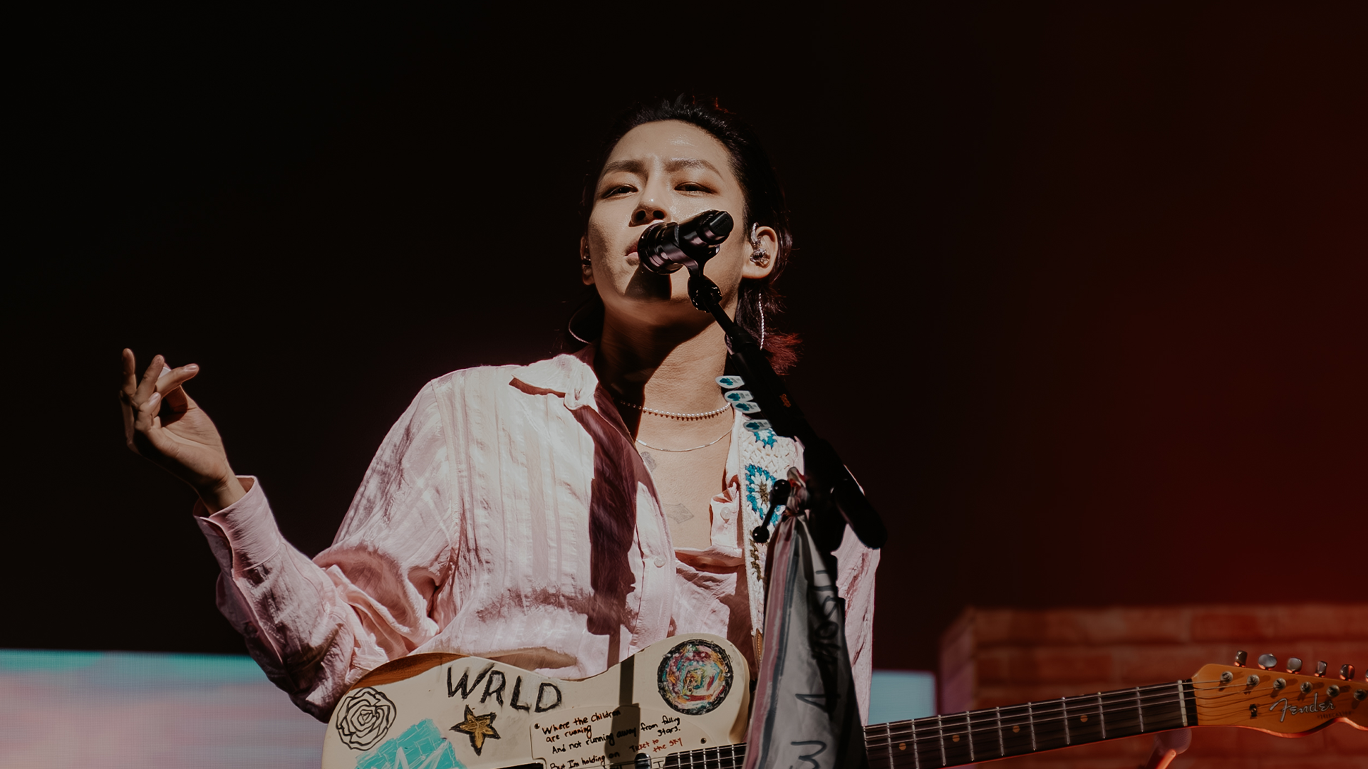 Woosung, vocalista do The Rose, cantando em um show ao vivo, segurando uma guitarra personalizada com desenhos e a inscrição WRLD, sob iluminação de palco.