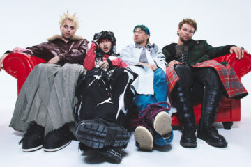 Banda 5 Seconds of Summer em ensaio promocional do álbum Everyone’s A Star! (2025), sentados em sofá vermelho com figurinos conceituais.