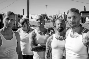 A banda Avenged Sevenfold posa com expressão séria, usando regatas brancas e exibindo tatuagens, em um cenário desértico.