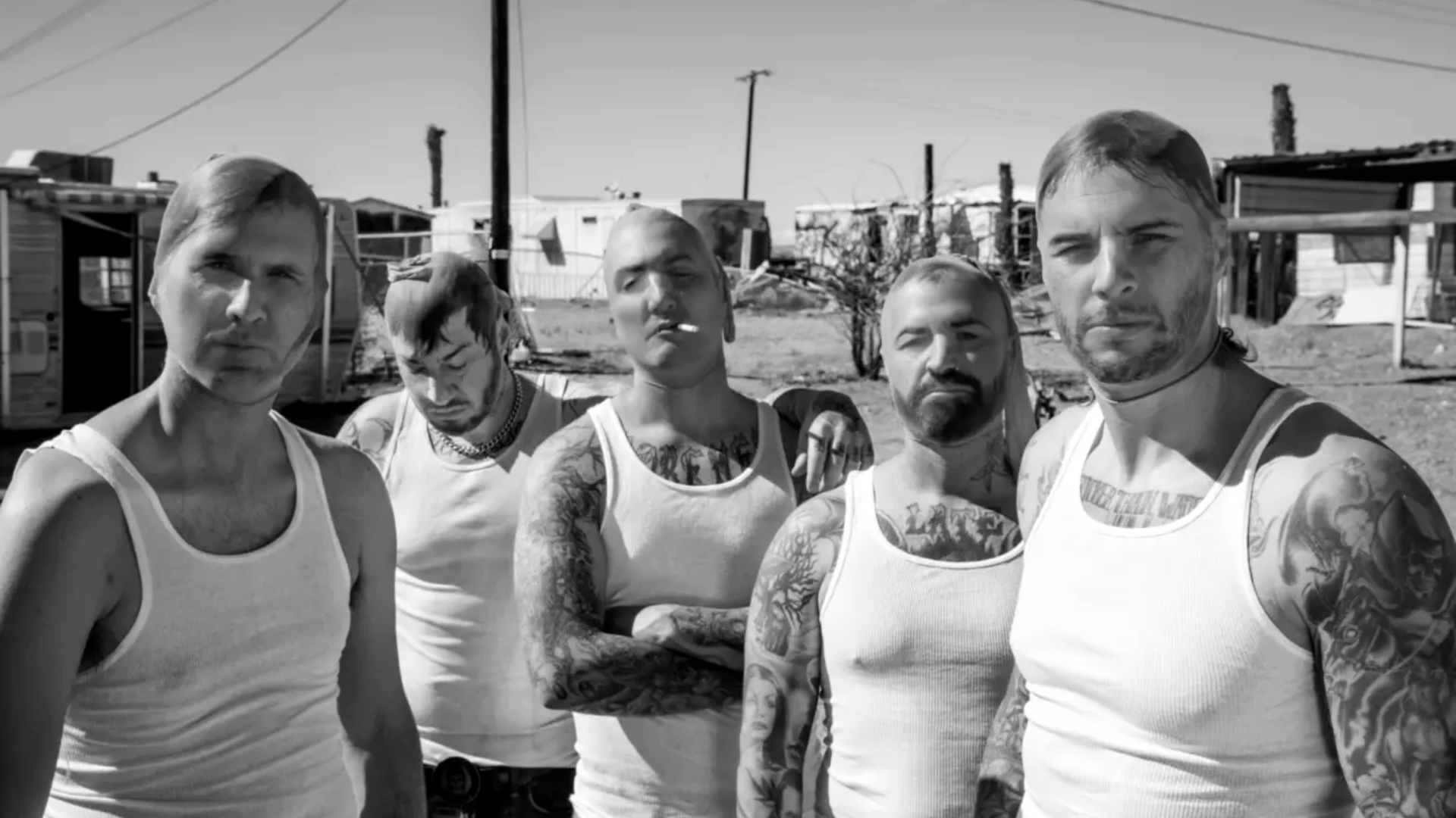 A banda Avenged Sevenfold posa com expressão séria, usando regatas brancas e exibindo tatuagens, em um cenário desértico.