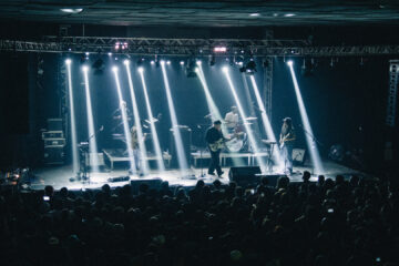 Terno Rei se apresentando no Tork n’ Roll em Curitiba com luzes verticais iluminando o palco e plateia lotada