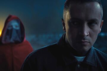Cena do videoclipe City Walls do Twenty One Pilots, com Tyler Joseph em destaque e uma figura encapuzada ao fundo.