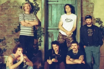 Integrantes do King Gizzard & The Lizard Wizard posam em frente a uma porta de madeira, com cenário de tijolos e plantas.