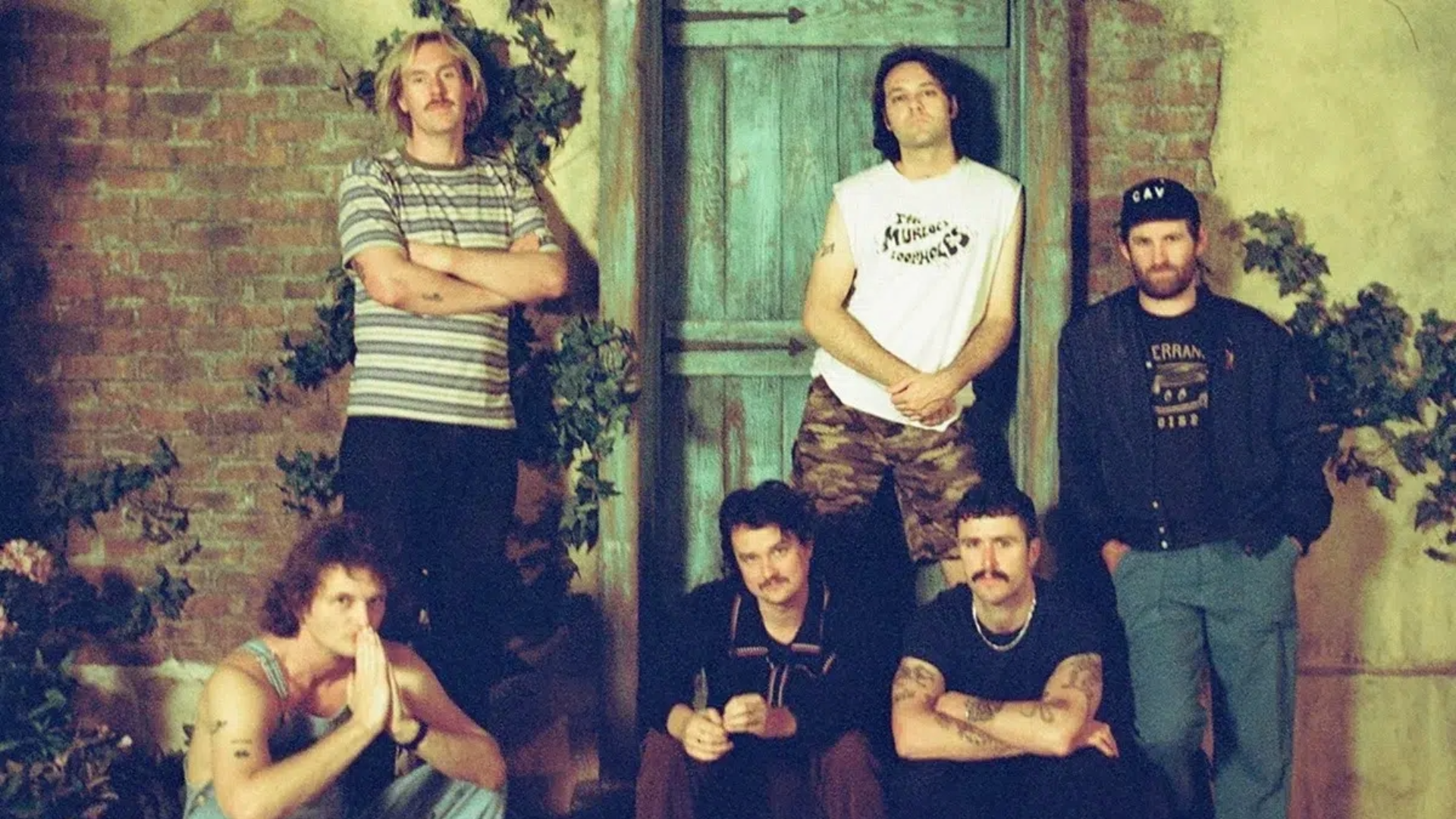 Integrantes do King Gizzard & The Lizard Wizard posam em frente a uma porta de madeira, com cenário de tijolos e plantas.