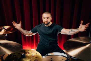 Eloy Casagrande, baterista do Slipknot, posando em seu kit de bateria com cortina vermelha ao fundo, em foto promocional tirada por Estevam Romera.