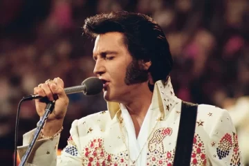 Elvis Presley se apresentando ao vivo em 1973 durante o especial Live in Honolulu, vestindo seu icônico macacão branco com pedras coloridas enquanto canta ao microfone.
