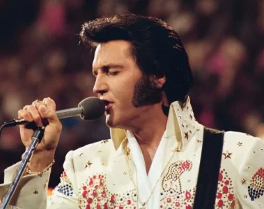 Elvis Presley se apresentando ao vivo em 1973 durante o especial Live in Honolulu, vestindo seu icônico macacão branco com pedras coloridas enquanto canta ao microfone.