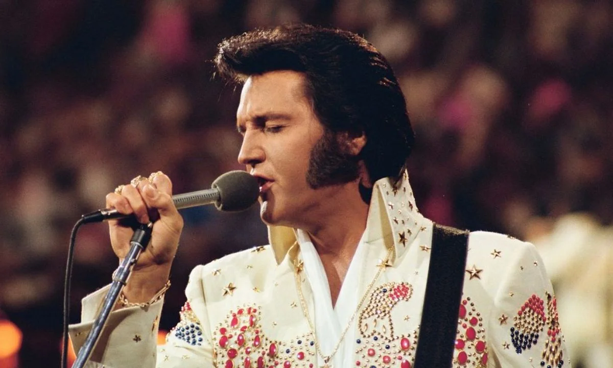 Elvis Presley se apresentando ao vivo em 1973 durante o especial Live in Honolulu, vestindo seu icônico macacão branco com pedras coloridas enquanto canta ao microfone.