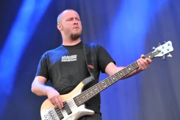 Sam Rivers tocando baixo durante show do Limp Bizkit, com expressão concentrada e fundo azul iluminado.