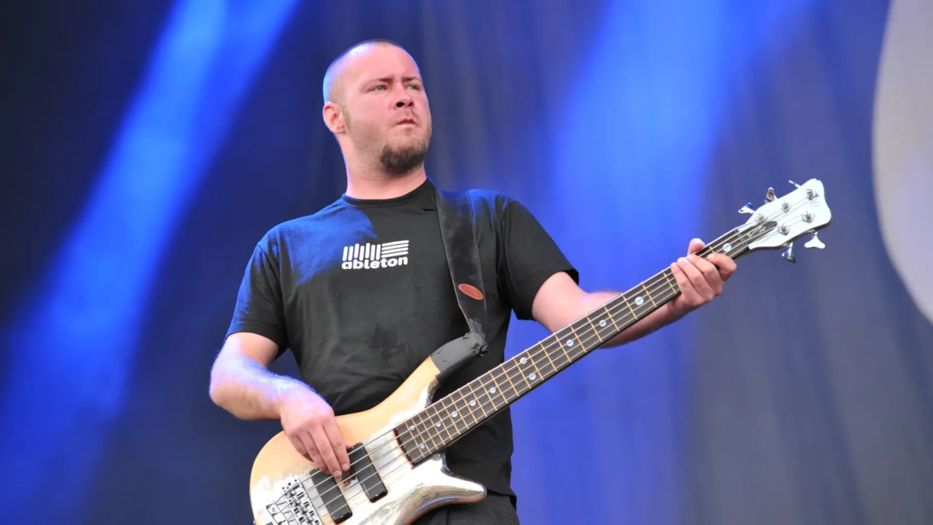 Sam Rivers tocando baixo durante show do Limp Bizkit, com expressão concentrada e fundo azul iluminado.