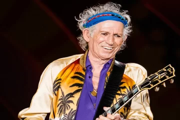 Keith Richards, guitarrista dos Rolling Stones, sorrindo durante uma apresentação no Estádio do Morumbi, em São Paulo, 2016, segurando uma guitarra Gibson e vestindo jaqueta dourada com estampa de palmeiras.